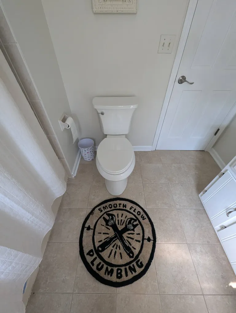 A toilet with a Smooth Flow branded rug on the bathroom floor.